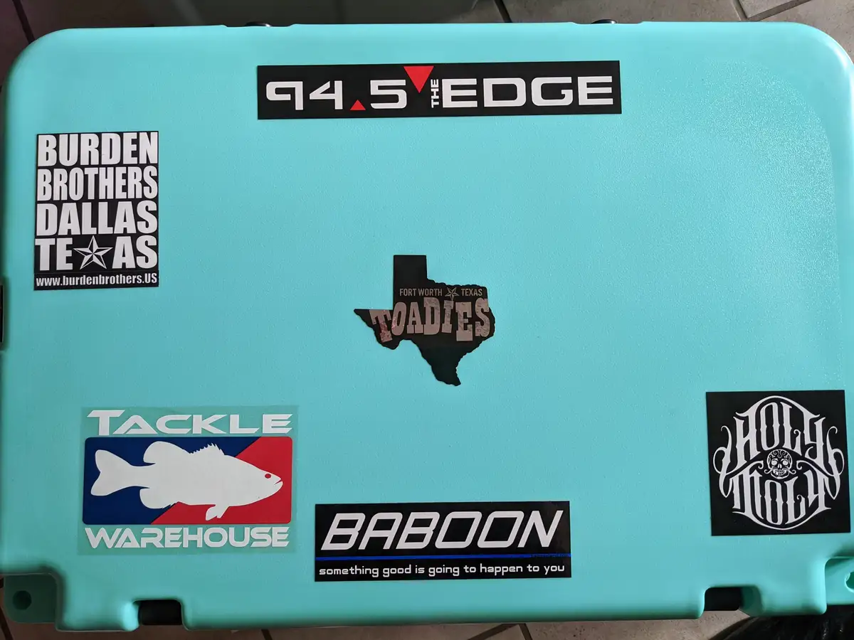 A light blue hard case is covered in various stickers including '94.5 THE EDGE', 'BURDEN BROTHERS DALLAS TEXAS', a…