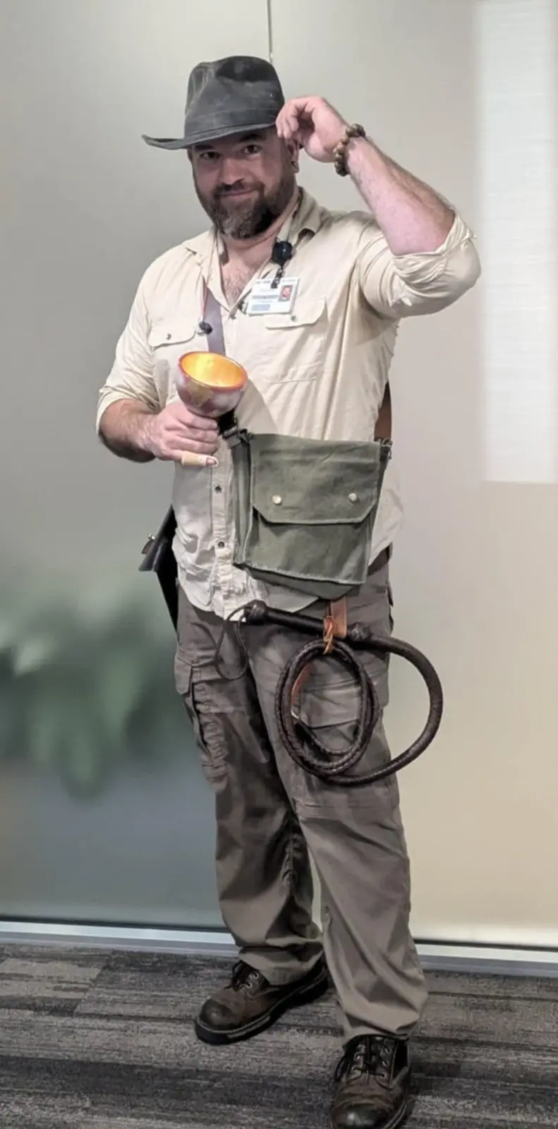 A man in an office setting, wearing a fedora, khaki shirt, cargo pants, and a work ID badge, poses with one hand on his hat…
