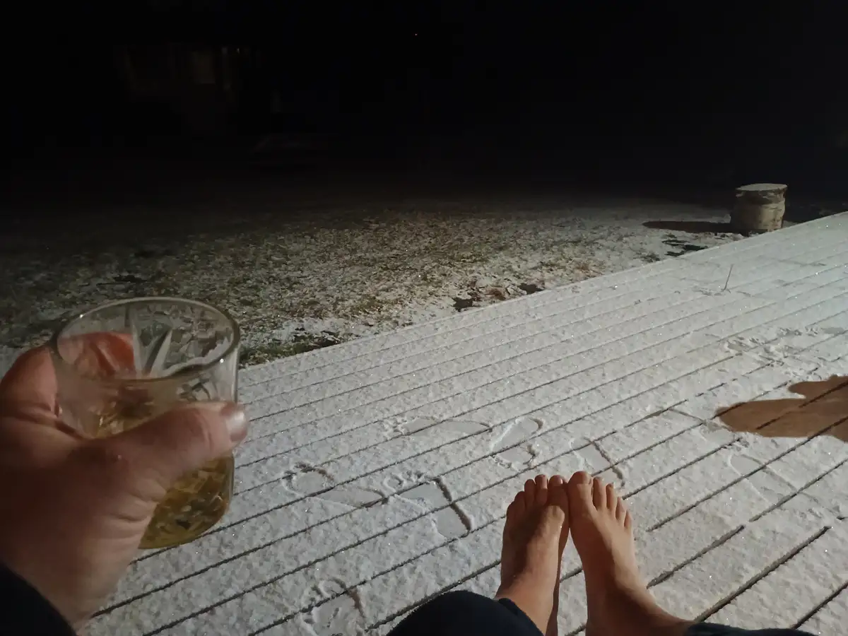 A first-person view shows bare feet on a frost-covered outdoor patio at night, with a hand holding a glass of amber liquid…