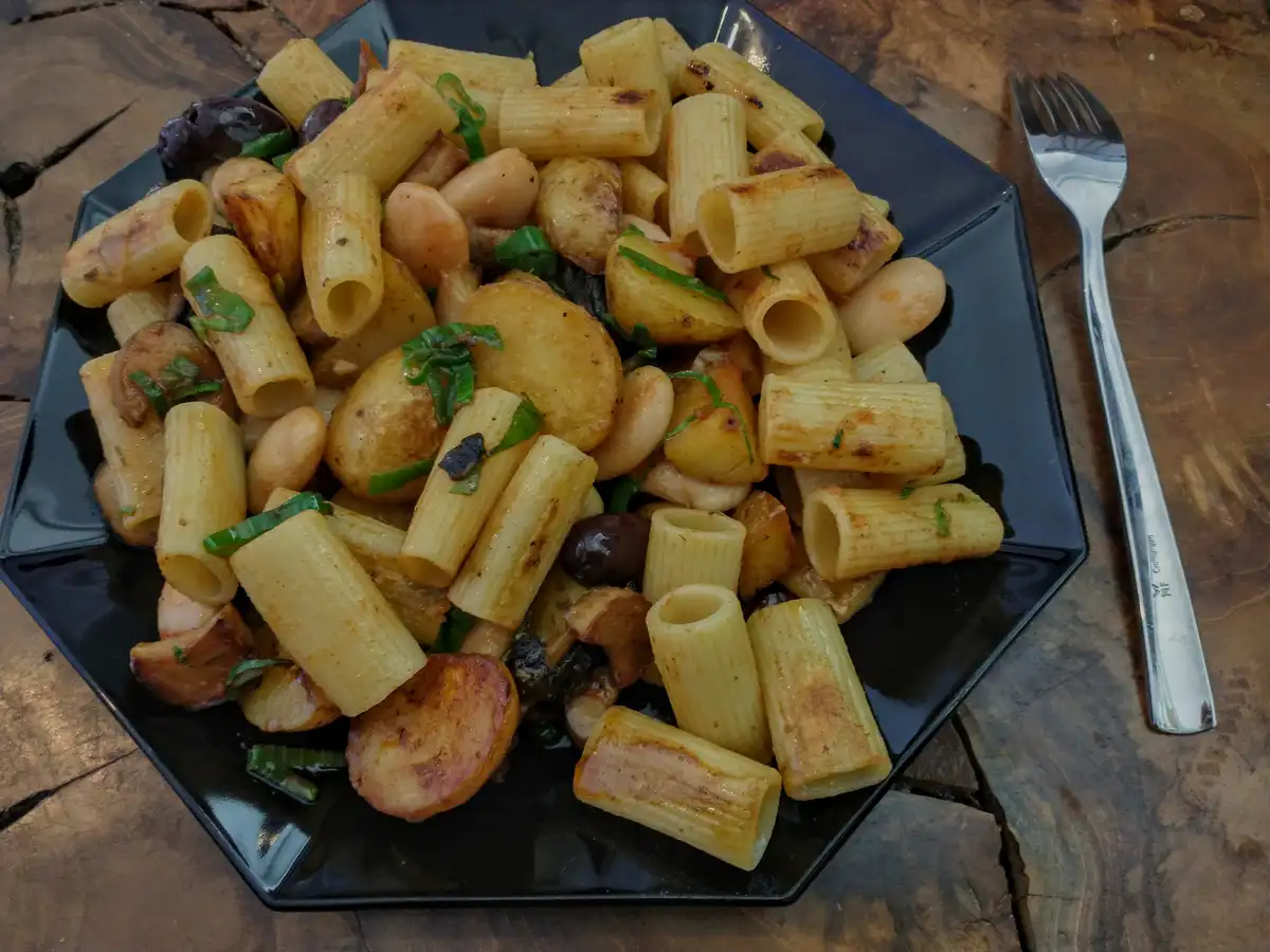 A black plate holds a dish of rigatoni pasta mixed with roasted potatoes, white beans, black olives, and chopped green…
