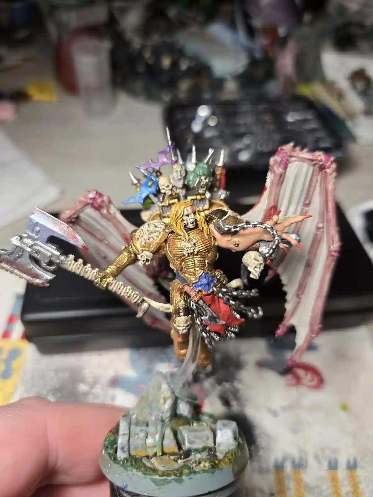 A highly detailed, painted miniature of a fantasy warrior with golden armor, skeletal wings, and a large axe, held by a…