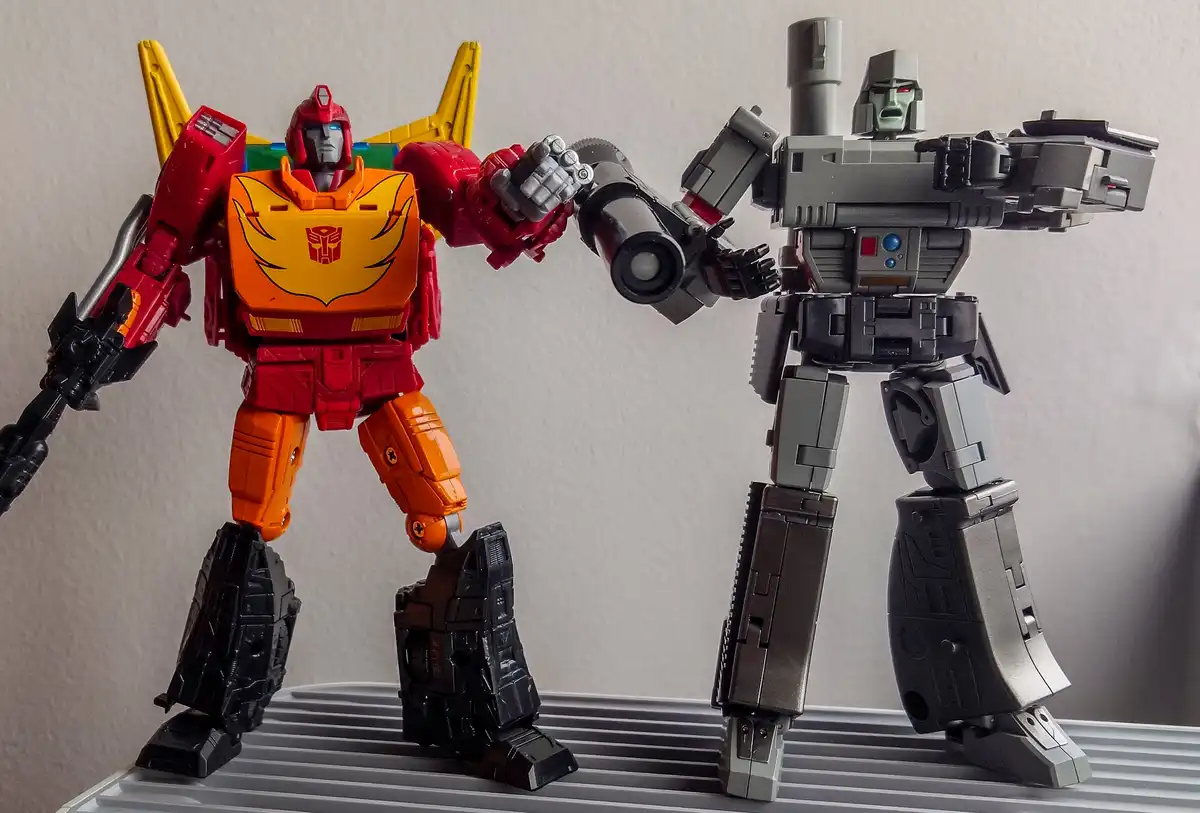 Two Transformers action figures stand facing each other, one red and orange with a yellow jetpack and Autobot symbol, the…