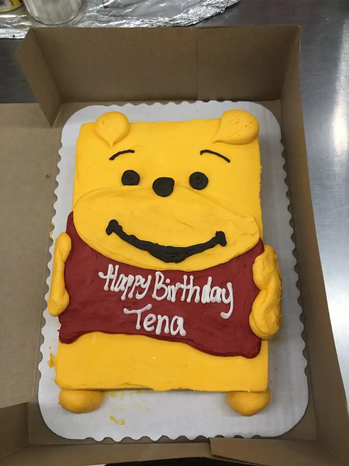 A rectangular birthday cake decorated to resemble Winnie the Pooh, with yellow frosting for his face and body, black icing…