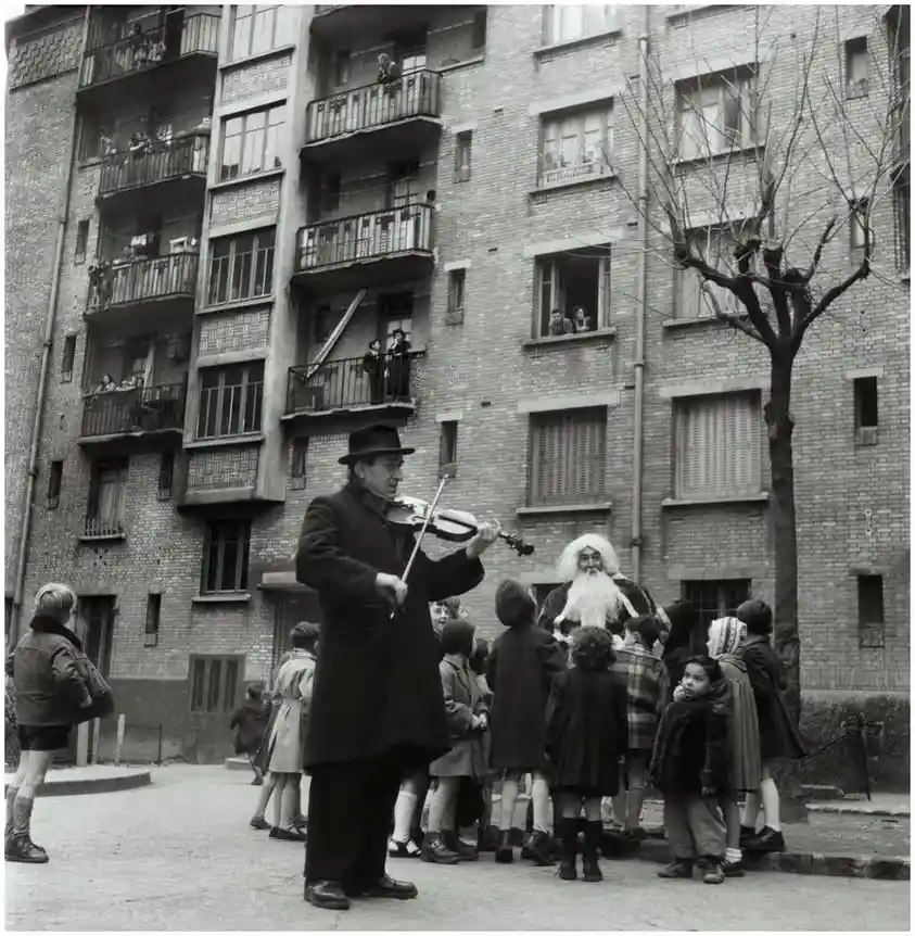 A black-and-white photo shows a man in a hat playing a violin for a group of children in a courtyard, while a man dressed as…