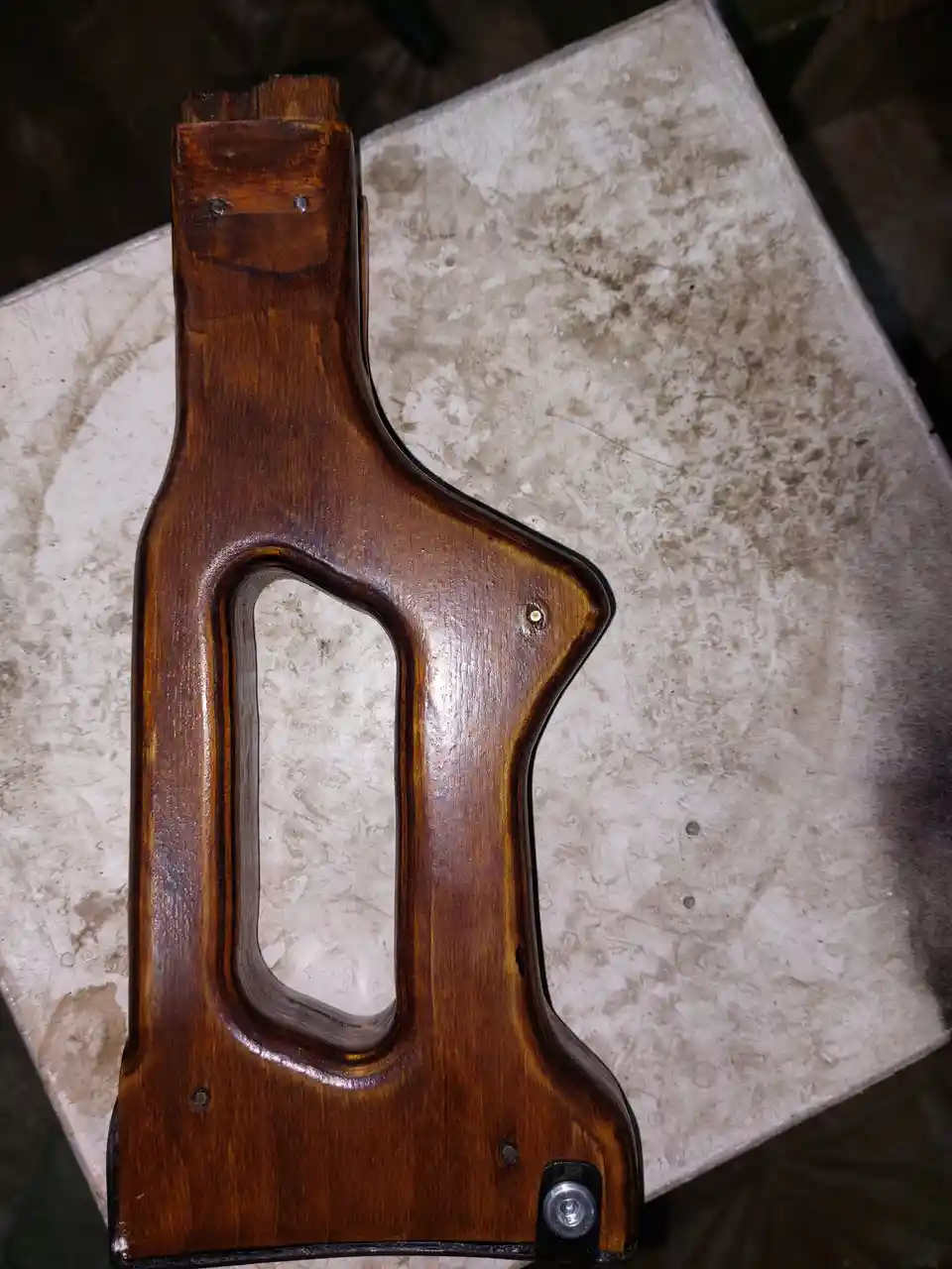 A close-up photo shows a wooden rifle stock with a glossy finish, resting on a worn, light-colored countertop. The stock has…