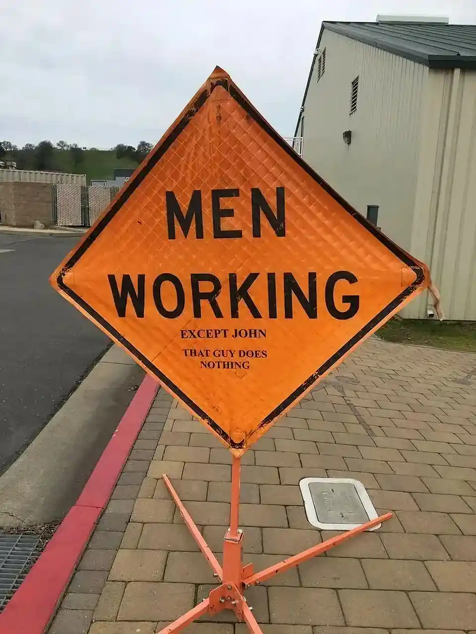 An orange diamond-shaped construction warning sign, typically reading 'MEN WORKING', has been altered with smaller text…