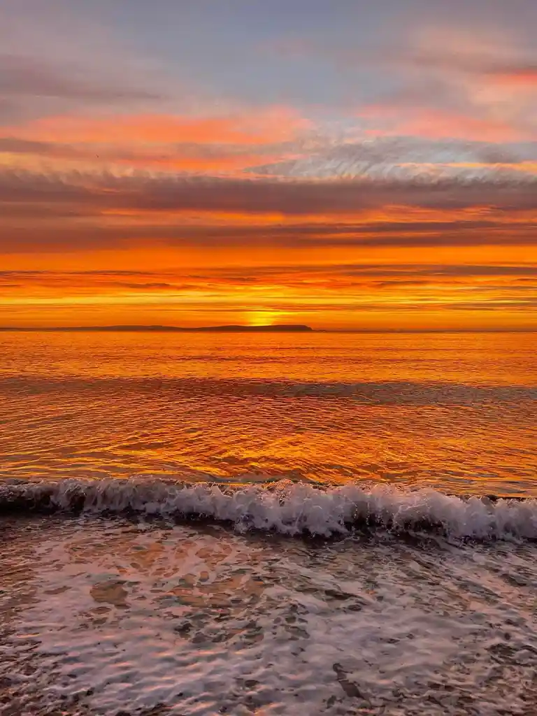 A serene sunset over the ocean with vibrant orange and pink clouds reflecting on the water, a small wave crashing onto the…