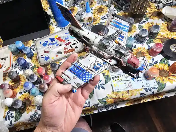 A hand holds a partially painted miniature spaceship model over a cluttered table covered in a sunflower-patterned cloth…