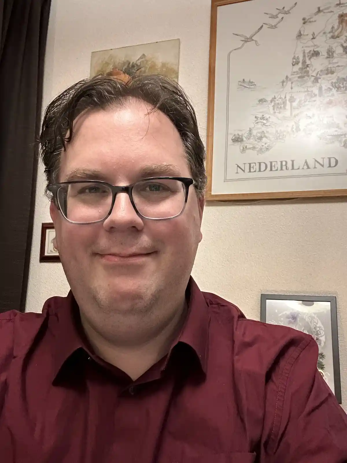 A man with dark hair and black-rimmed glasses, wearing a maroon collared shirt, smiles slightly at the camera. Behind him on…