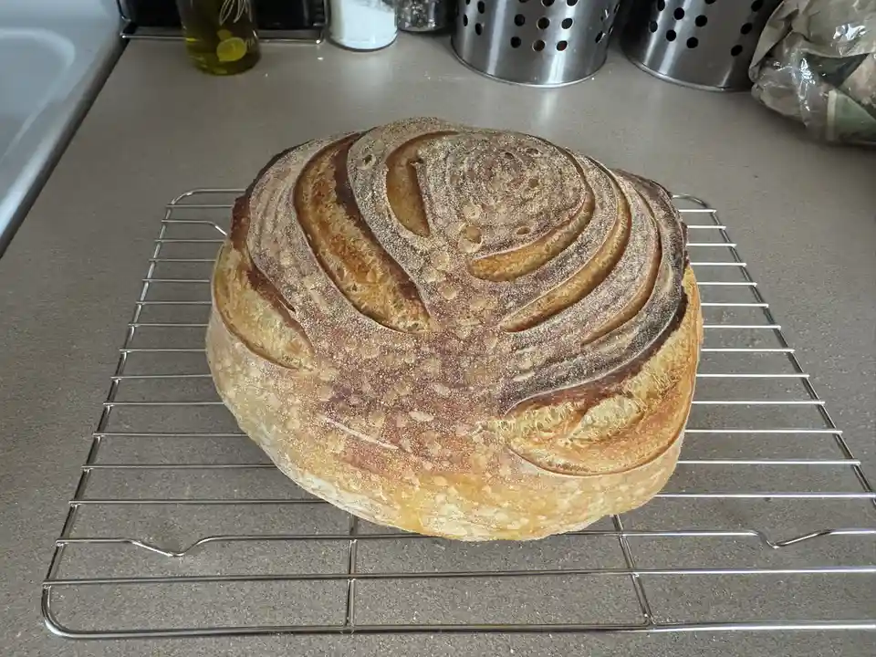 A round, golden-brown loaf of artisan bread with a scored top sits on a metal cooling rack on a kitchen counter. The scoring…