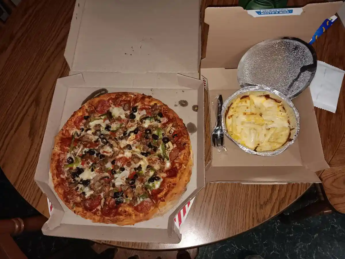 An overhead photo shows a large, open pizza box with a loaded pizza featuring pepperoni, olives, sausage, and peppers…