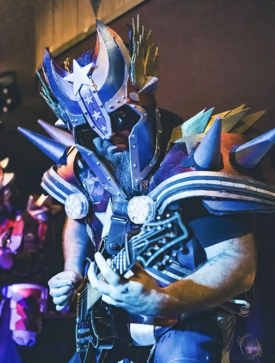 A man with a full beard wears an elaborate, heavily armored costume with spikes, stars, and glowing lights, playing a…