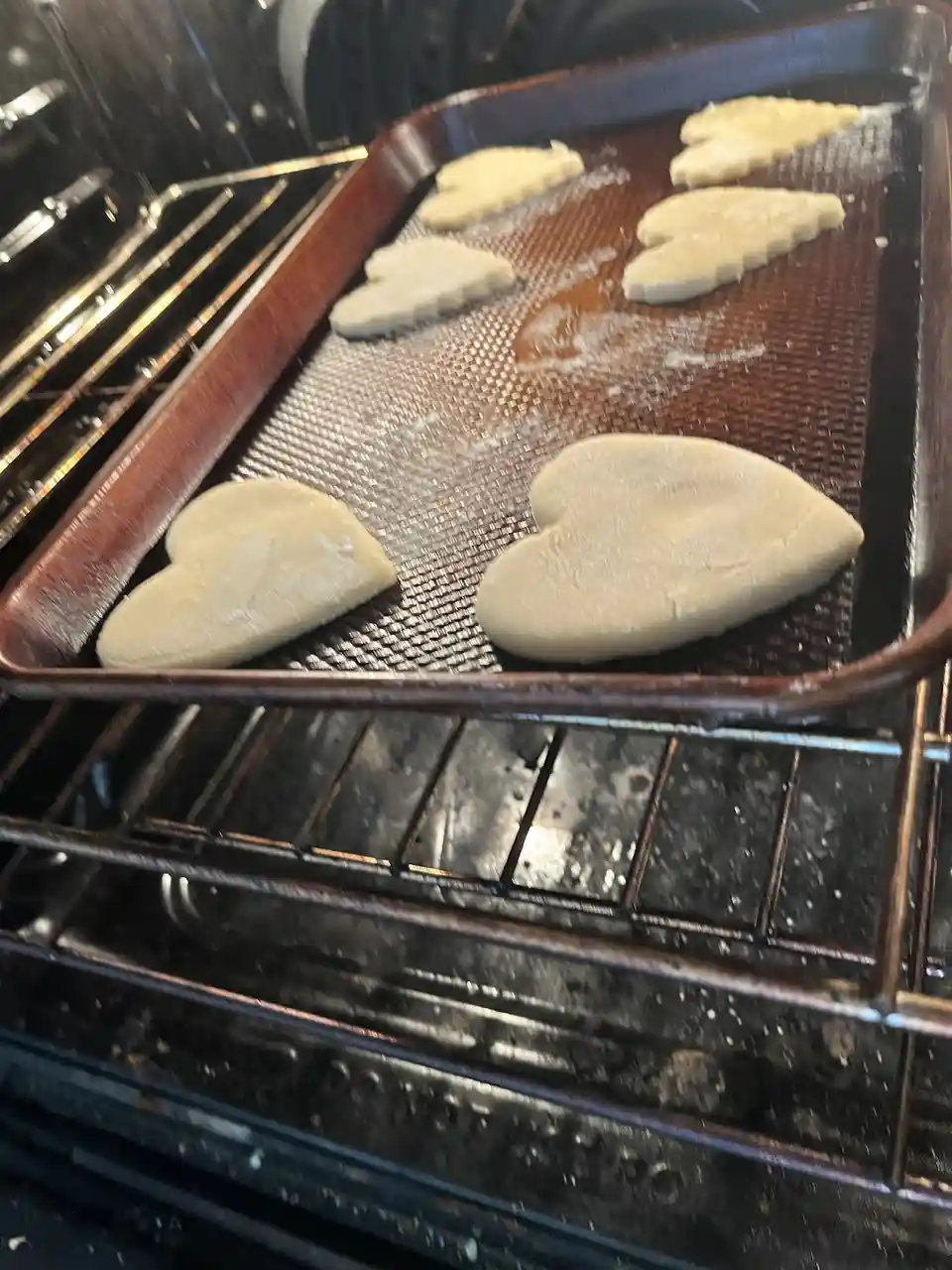 A close-up photo shows a baking tray inside an oven, holding several heart-shaped cookies made from pale dough. The cookies…