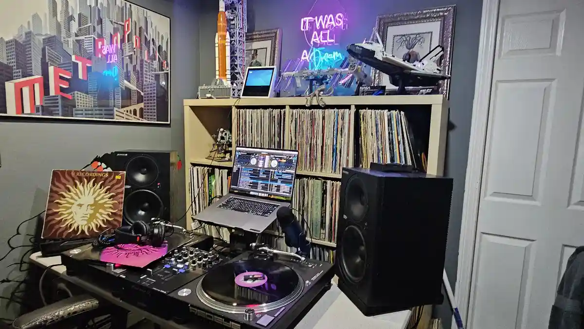 A room with a DJ setup including turntables, a laptop running DJ software, and large speakers, surrounded by shelves of…