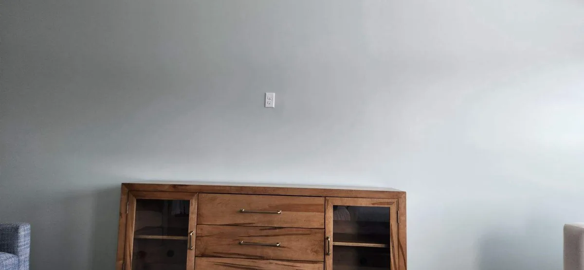 A plain photo shows a white electrical outlet mounted high on a light gray wall, directly above a wooden media console with…