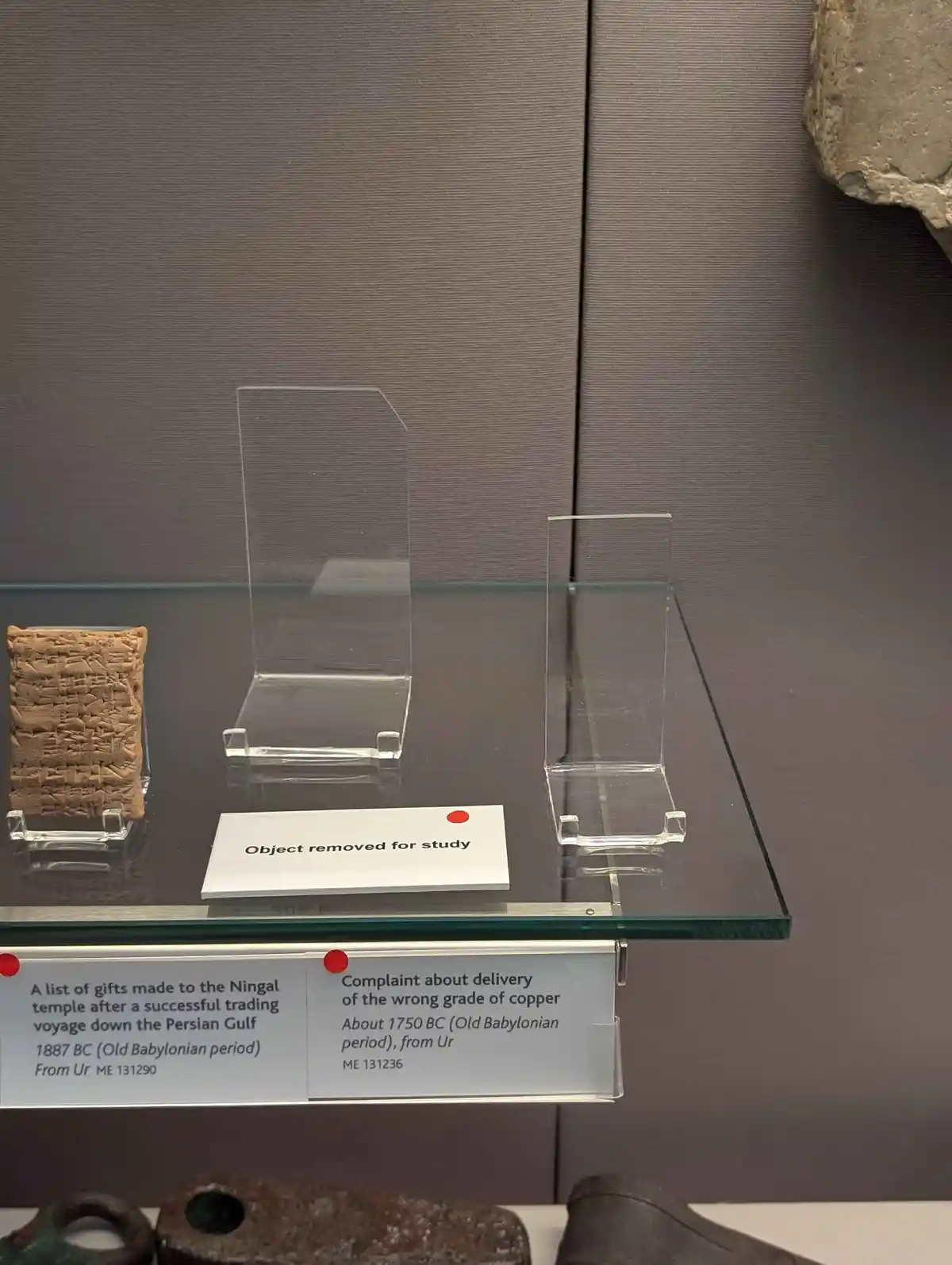A museum display case holds two ancient cuneiform tablets on clear acrylic stands, with a white card in the center stating…