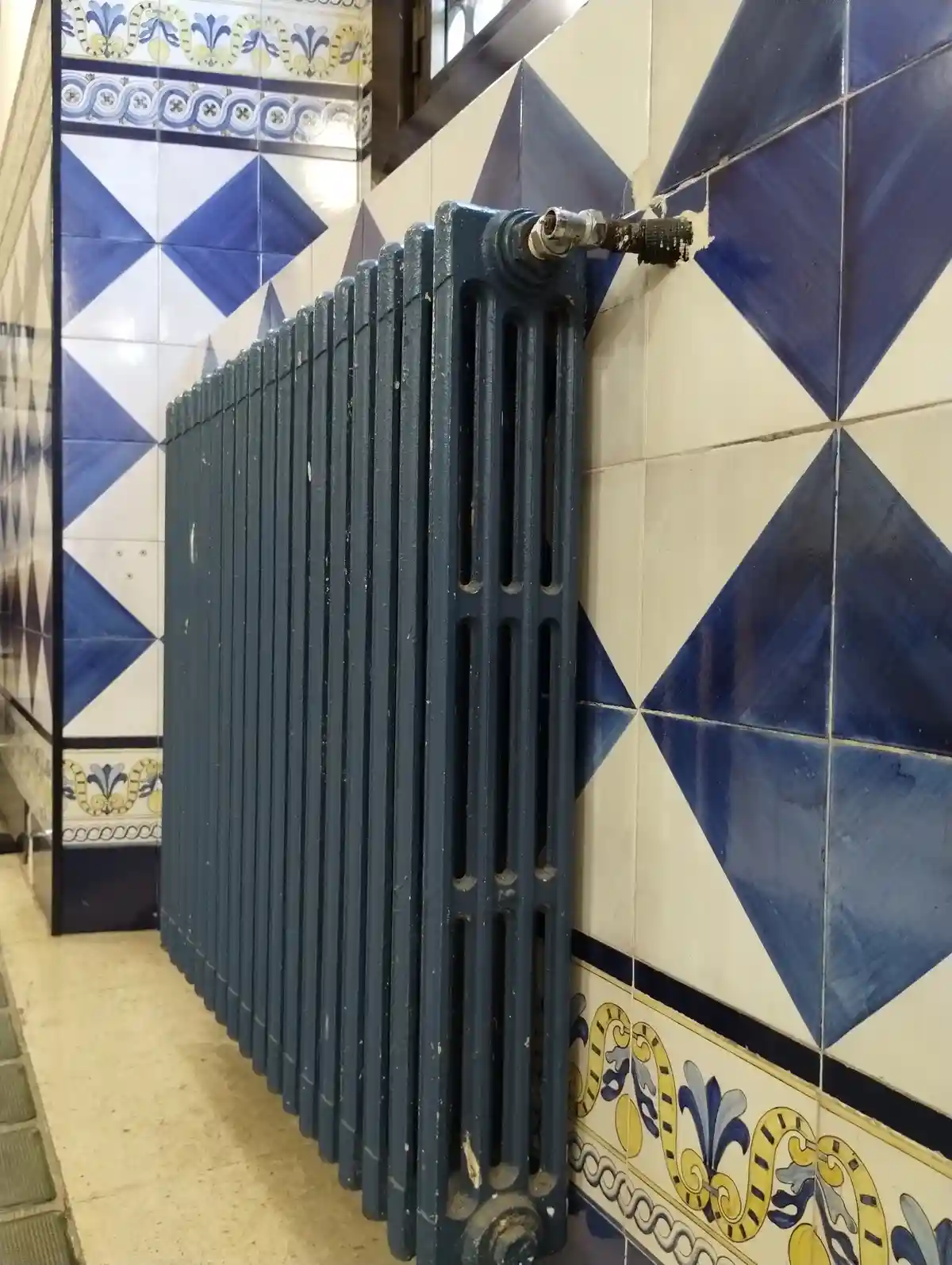 A dark blue, vintage-style radiator is mounted against a wall covered in ornate blue and white geometric tiles with…