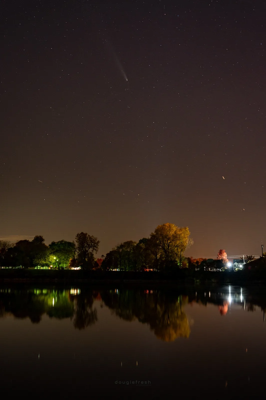 A night sky photograph shows a starry sky with a visible comet tail and faint airplane contrails, above a treeline and calm…