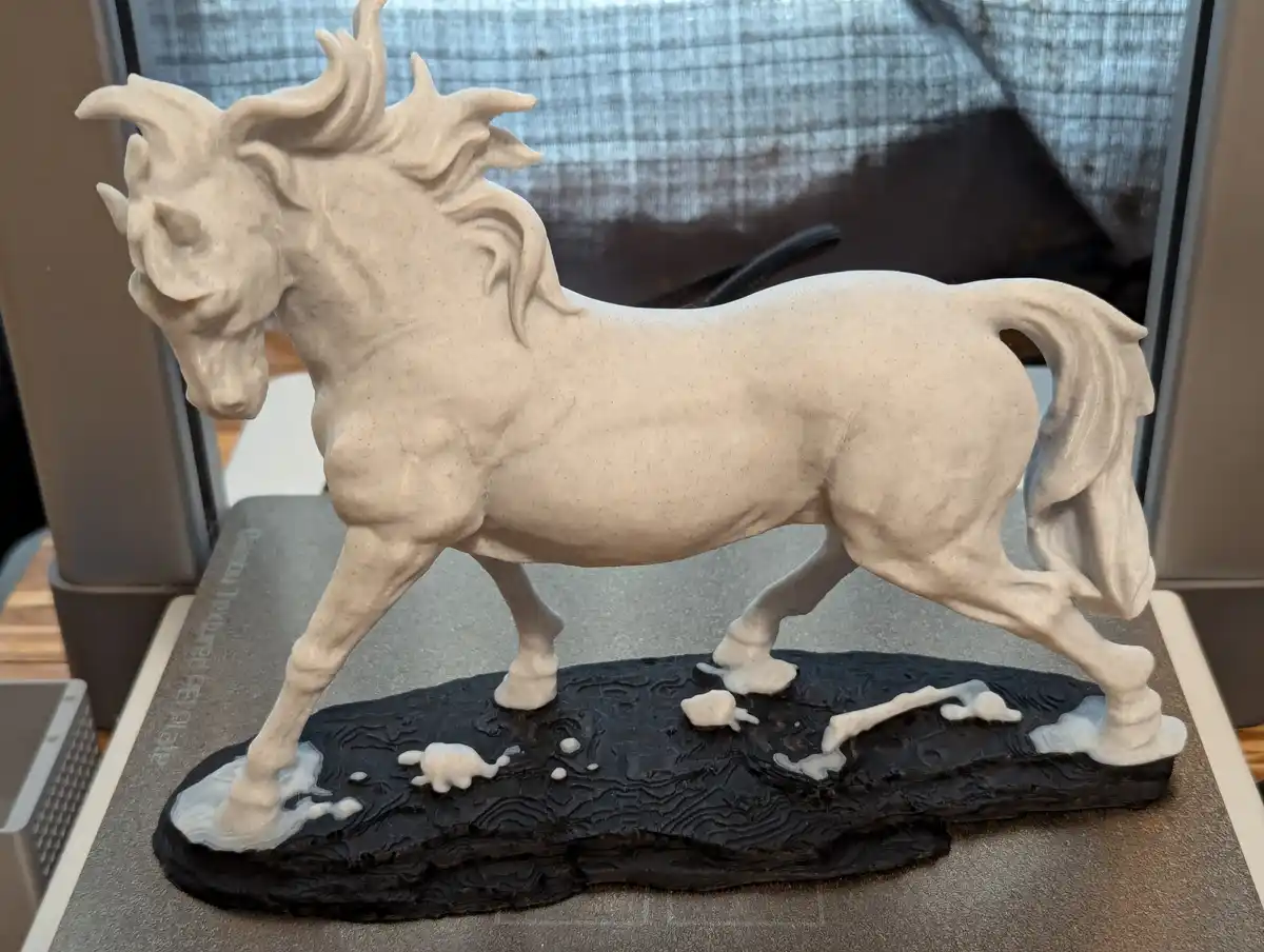 A white horse statue with a flowing mane and tail stands on a black base resembling water or mud, with white splashes around…