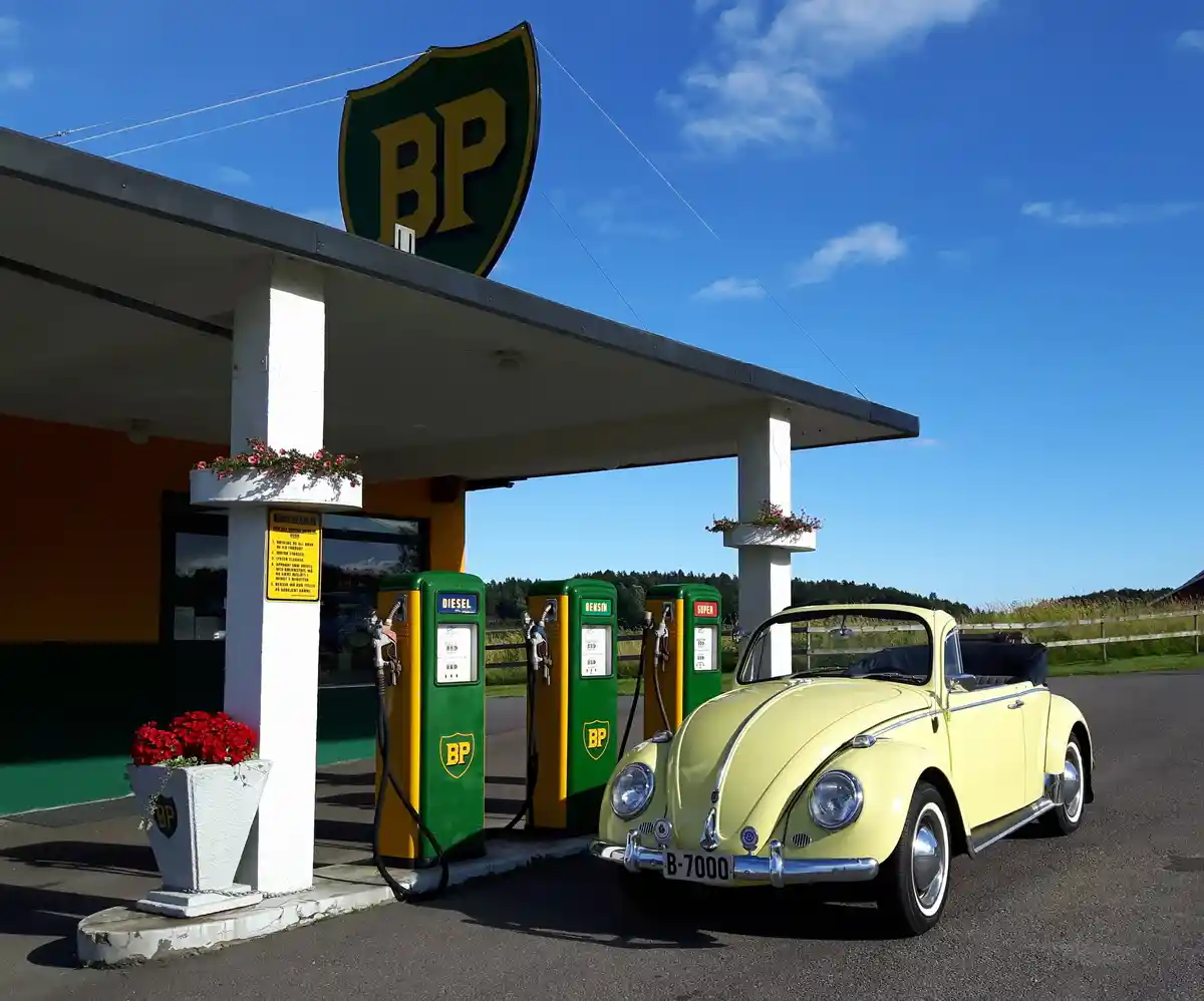 A vintage yellow Volkswagen Beetle convertible is parked at a retro-style BP gas station under a clear blue sky. The station…