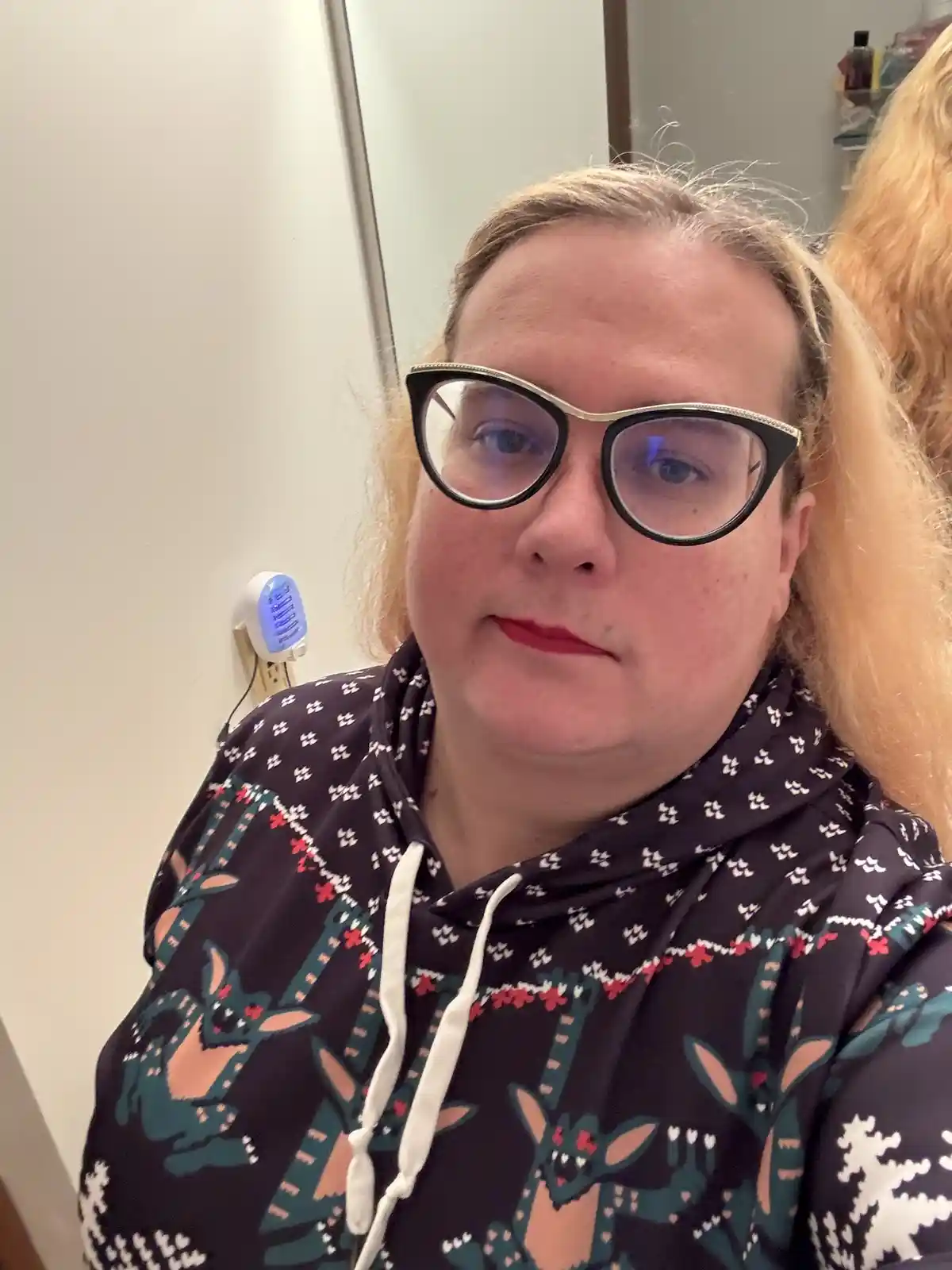 A person with blonde hair and black-rimmed glasses wearing a dark hoodie with a festive pattern of reindeer and snowflakes…