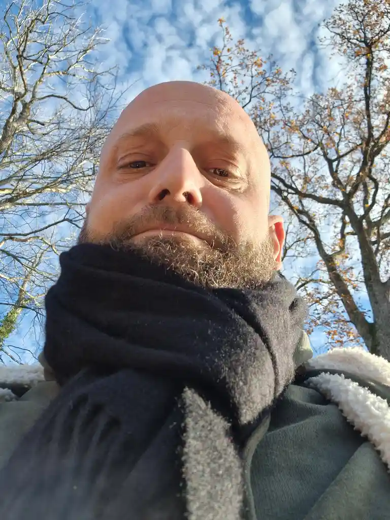 A low-angle selfie of a bald man with a beard, wearing a dark scarf and a jacket with a white fleece collar, outdoors under…