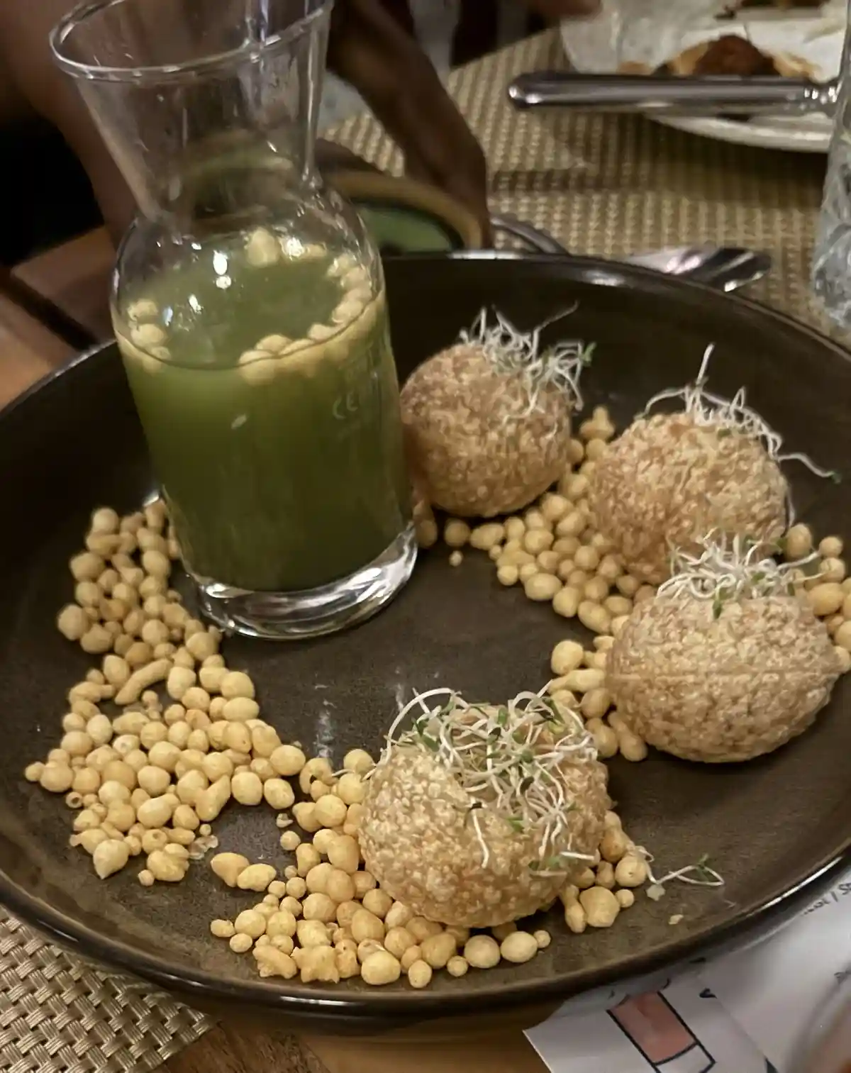 A dark plate holds four round, textured fried balls topped with sprouts, surrounded by small golden fried snacks, next to a…