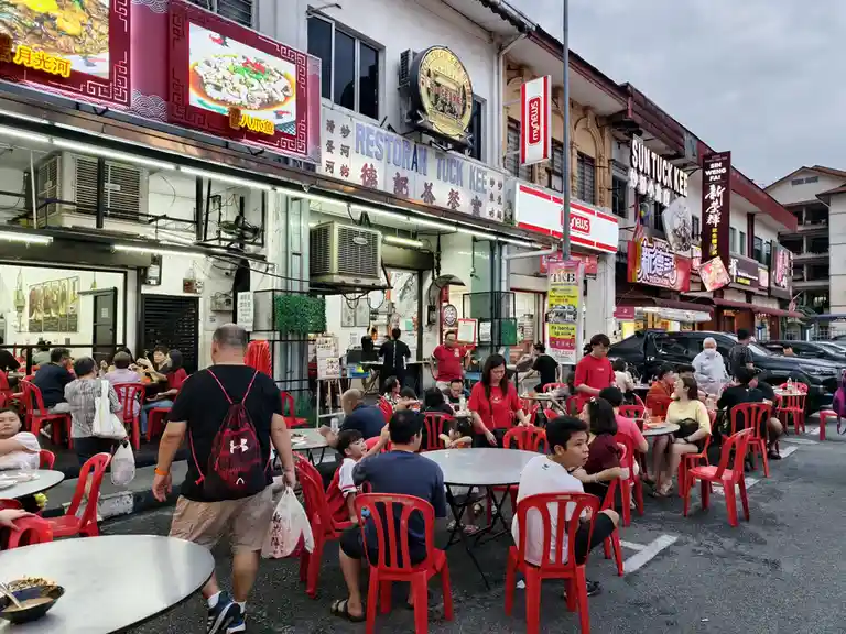 A bustling street food scene in Malaysia with many people seated at red plastic tables and chairs outside a restaurant named…