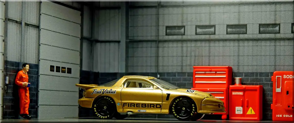 A miniature gold Pontiac Firebird race car with 'True Value' and 'Firebird' decals is parked in a garage next to a miniature…
