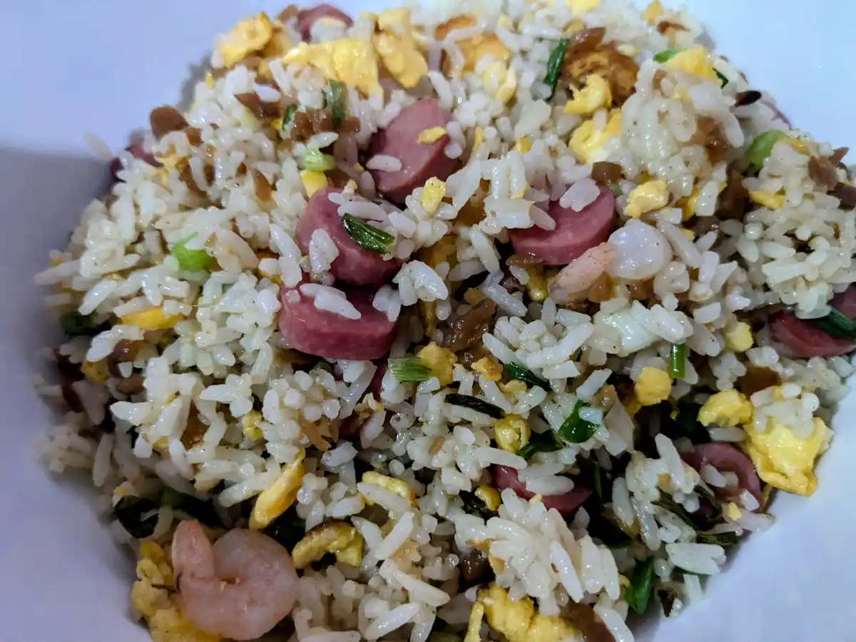 a close-up photo of a white bowl filled with fried rice containing visible ingredients like scrambled egg, chopped green…