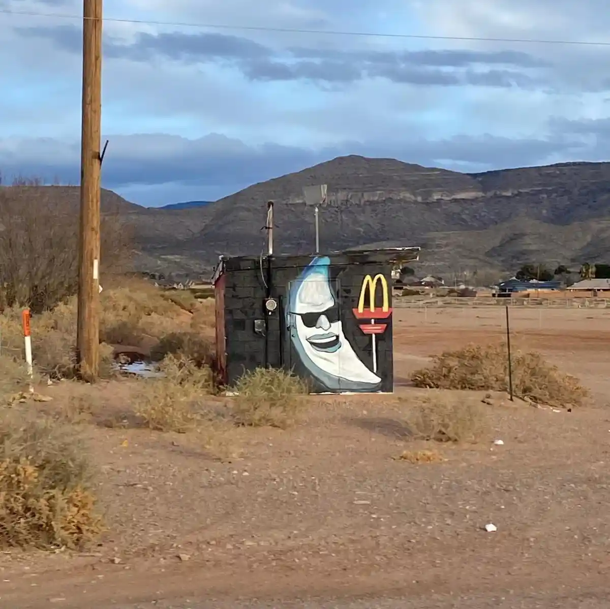 A desert landscape features a small, weathered utility box painted with a stylized crescent moon wearing sunglasses and…