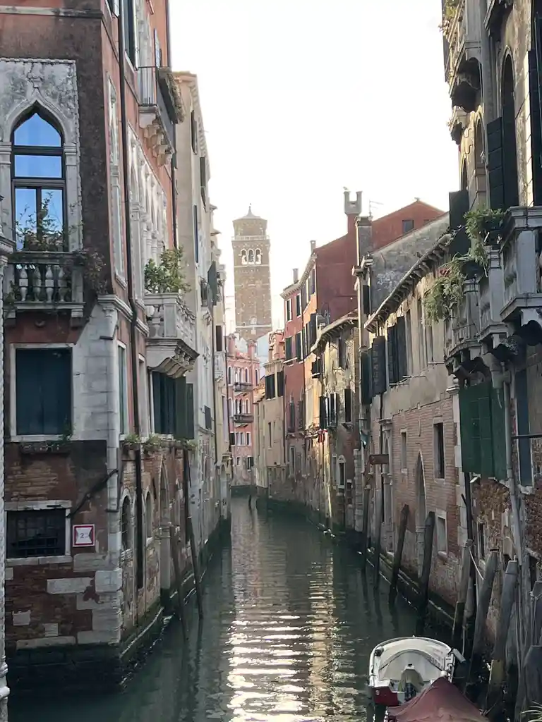 A narrow Venetian canal flanked by historic, colorful buildings with ornate balconies and shuttered windows, leading toward…