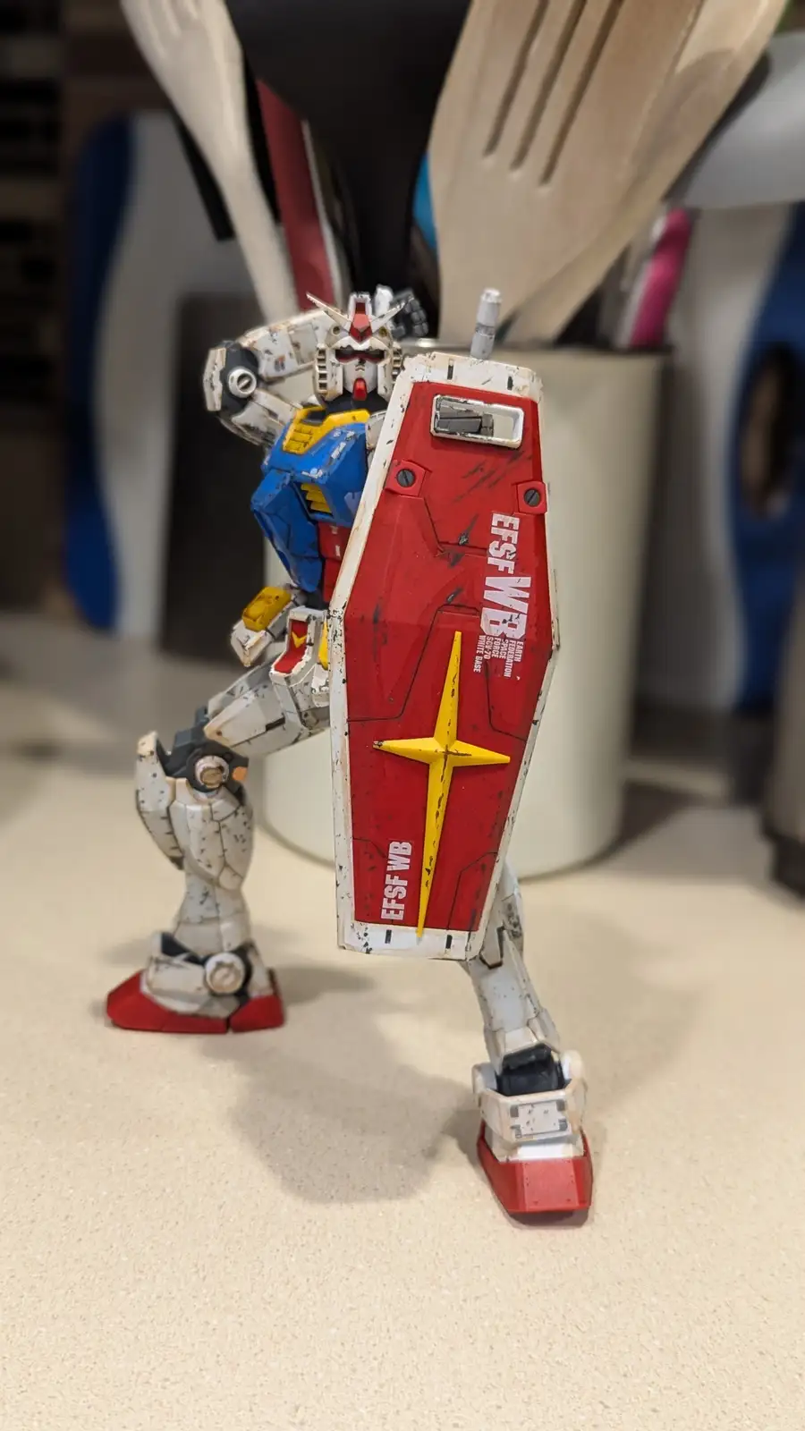 A weathered, white, blue, and red Gundam model stands on a kitchen countertop holding a red shield with a yellow star and…