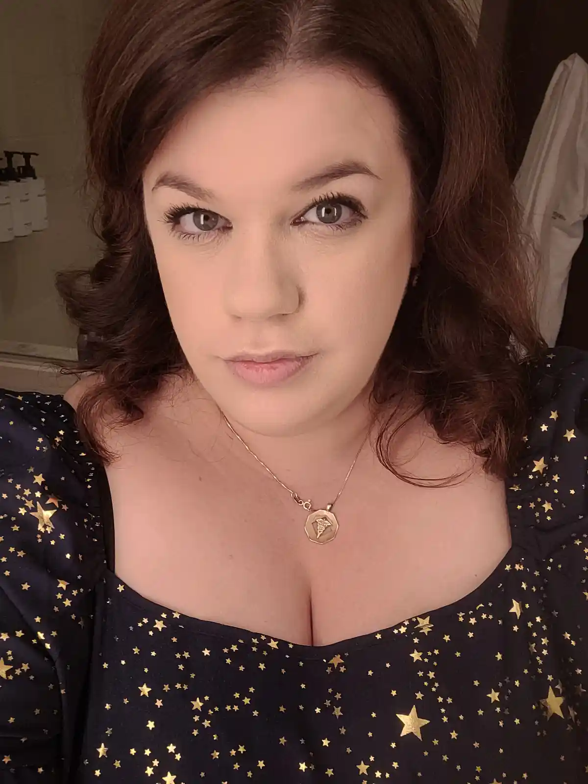 A woman with brown hair and light eyes stares directly at the camera with a serious expression. She wears a black top with…