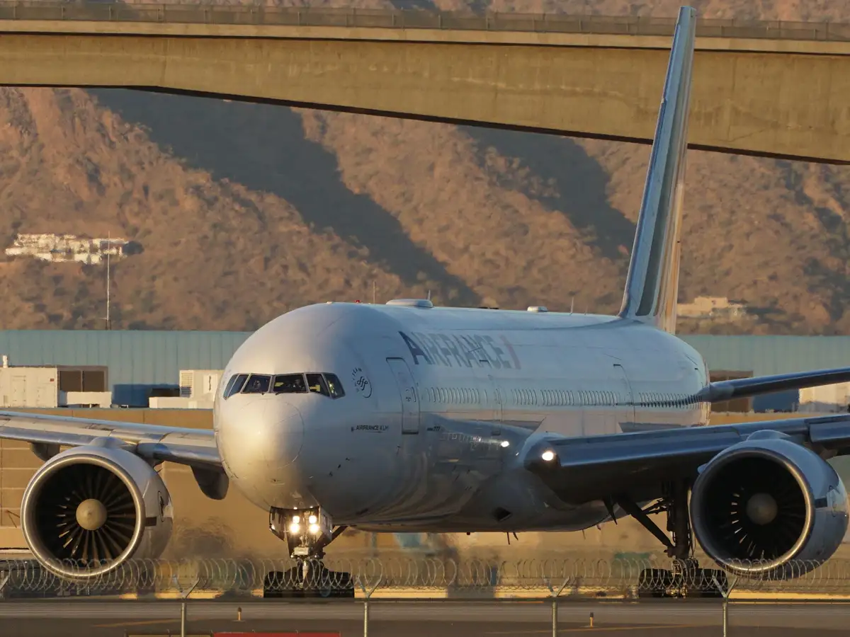 A large Air France Boeing 777 airplane is parked on an airport tarmac under a concrete overpass, with mountains in the…