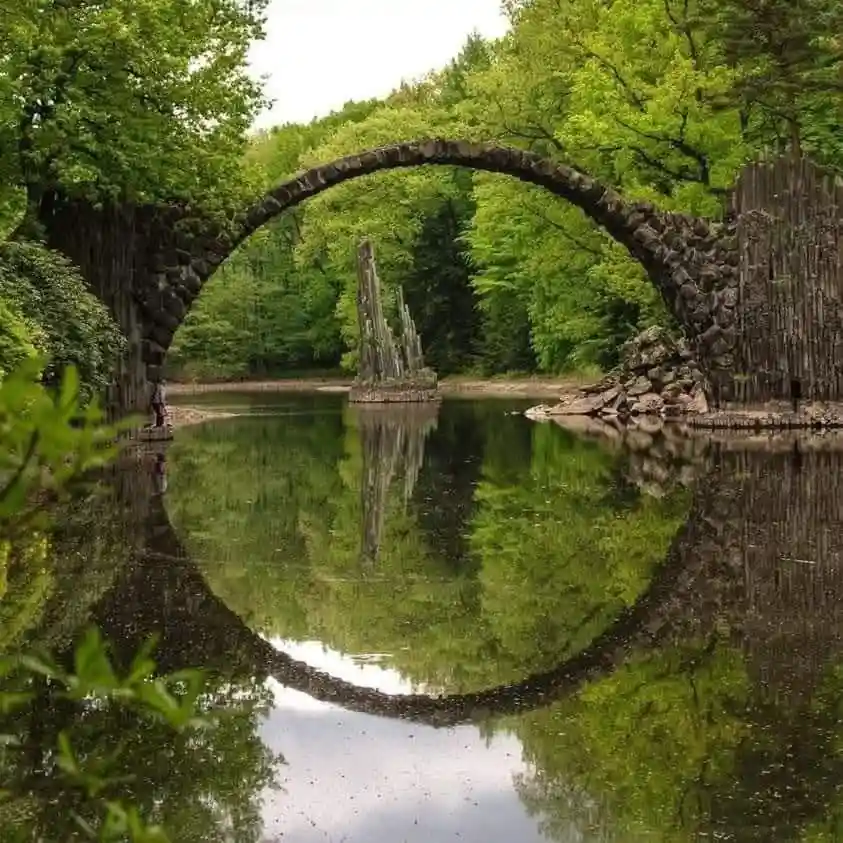 a natural stone arch bridge over a calm, reflective pond in a lush green forest, perfectly mirrored in the water to form a…