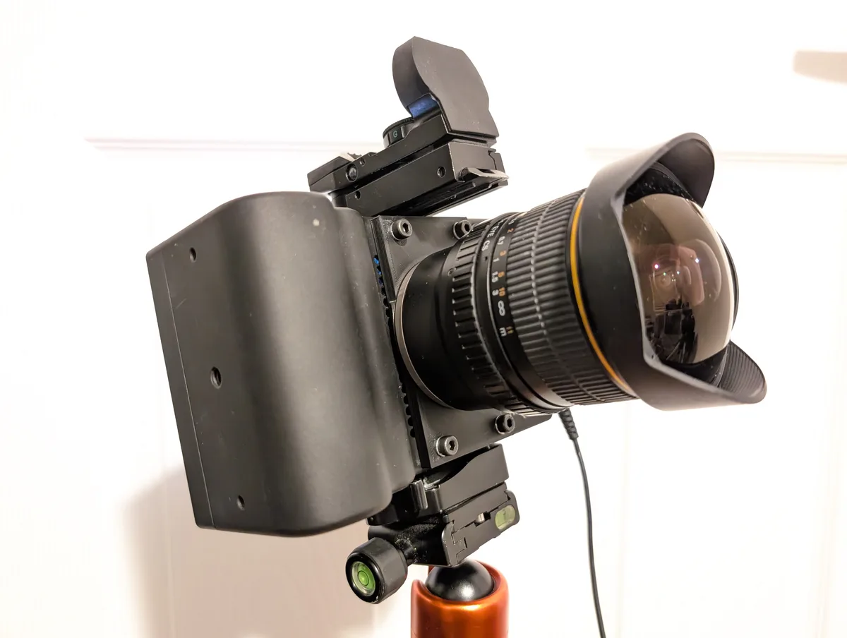 A black DSLR camera with a wide-angle fisheye lens and a custom black housing is mounted on a tripod against a white wall…