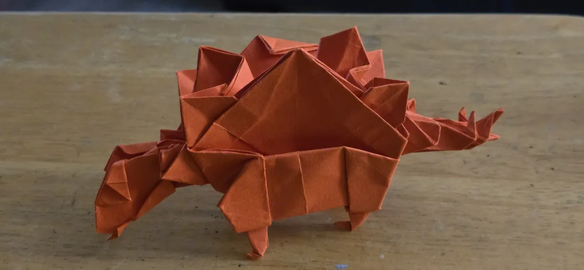 An orange origami dinosaur with a spiked back and tail sits on a wooden table. The overlaid text reads 'i made this origami…