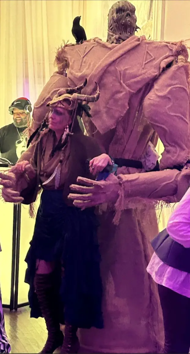 A person in a large, monstrous costume with a bird perched on their shoulder stands beside a woman in a fantasy-themed…