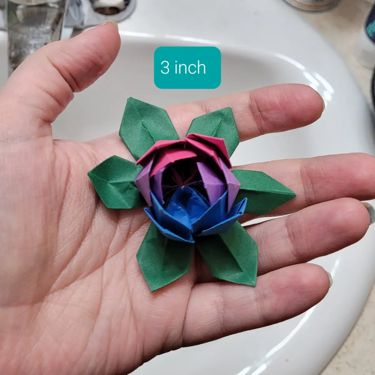 A hand holds a small, multi-colored origami flower with green leaves, positioned over a white sink. A teal text box above…