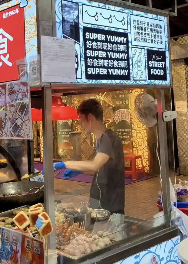 A street food vendor in a glass-fronted stall is cooking, wearing blue gloves and earphones, with a large sign above him…