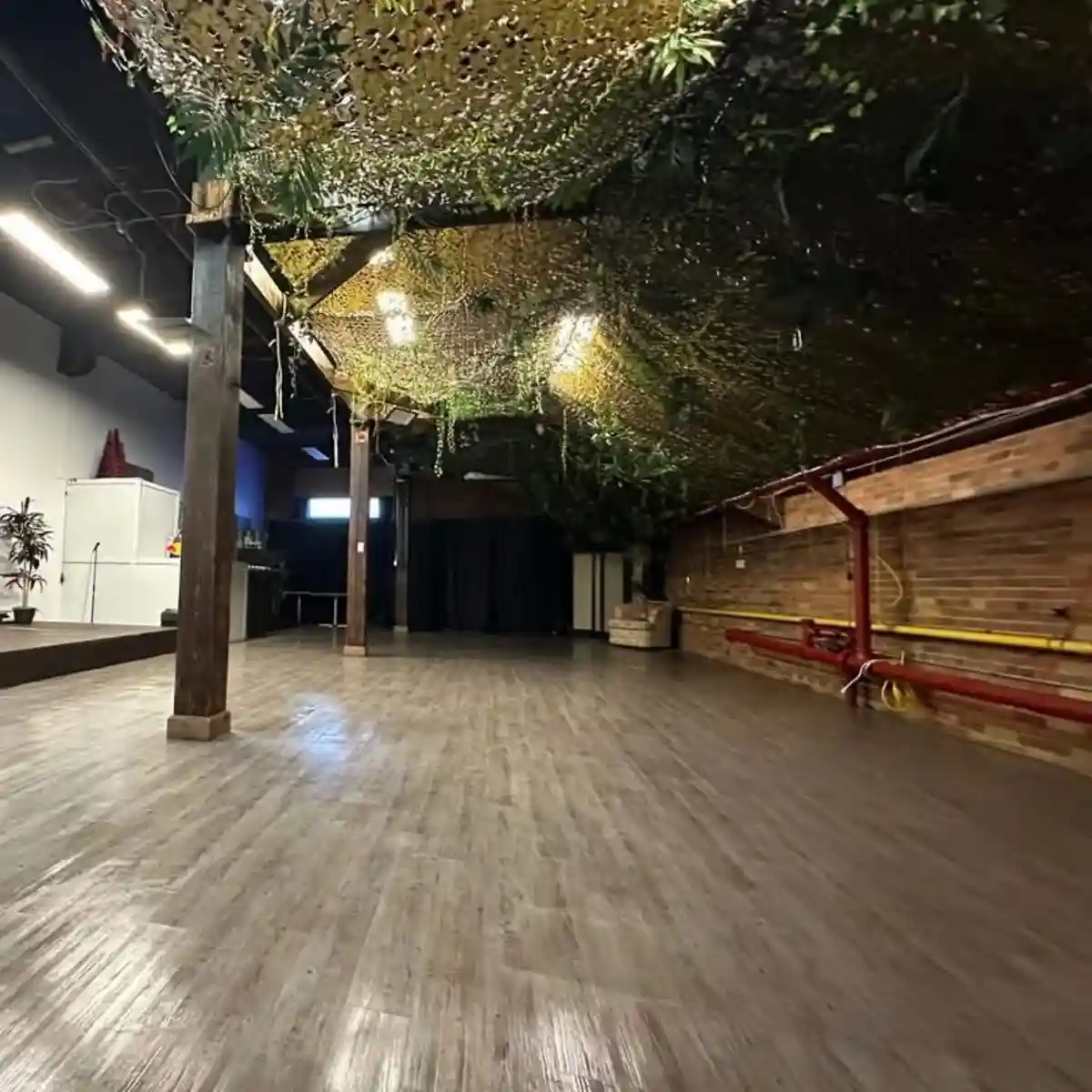 An empty dance studio with wooden floors, exposed beams, and brick walls, but the ceiling is covered in dense artificial…