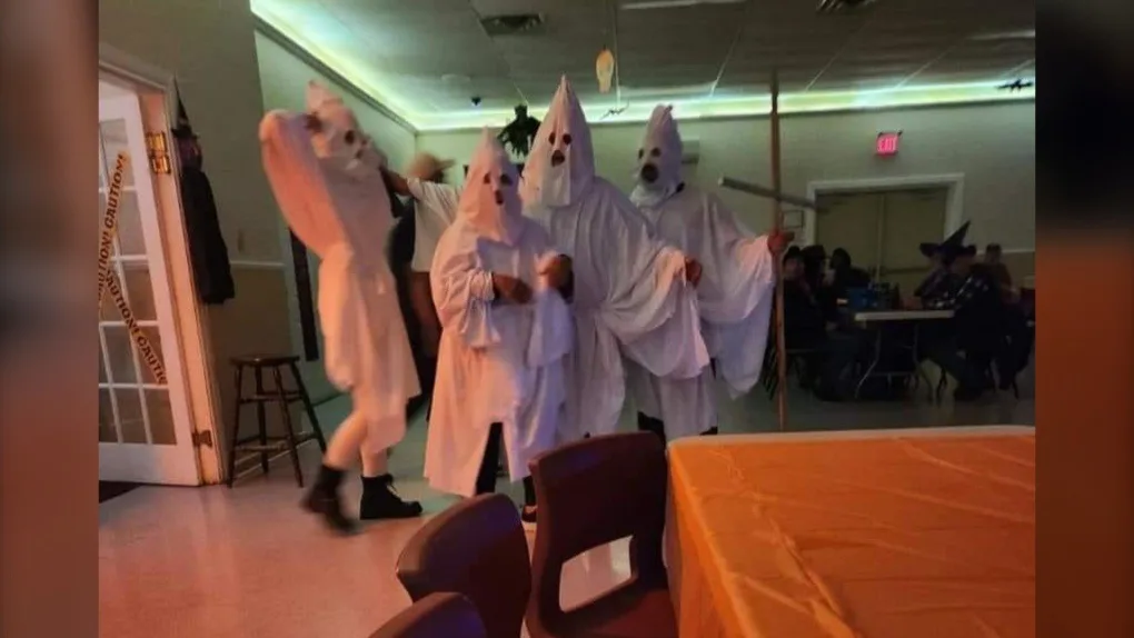 Several people in white ghost costumes with cone-shaped hoods are dancing in a room; one is mid-motion with arms raised…