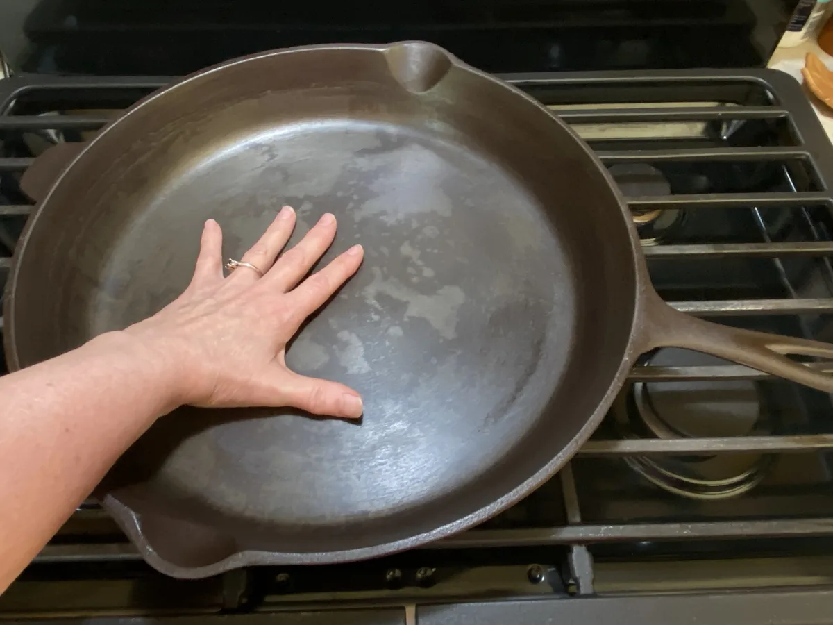 A hand with a ring rests flat inside a large, well-seasoned cast iron skillet sitting on a gas stove grate. The pan's…