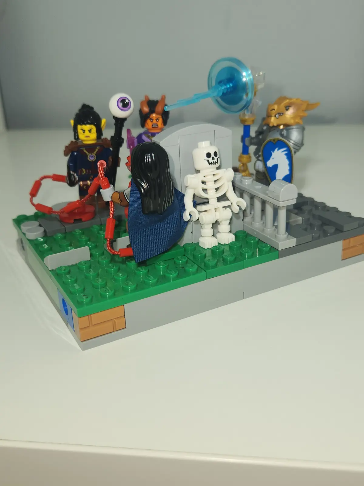 A LEGO diorama shows a skeleton standing in front of a tombstone, with a figure in a blue robe facing away from the viewer…