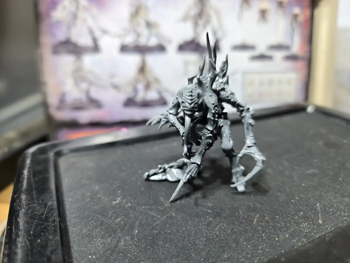 A gray, unpainted miniature of a spiky, monstrous creature with multiple limbs and a spiked head stands on a scratched black…