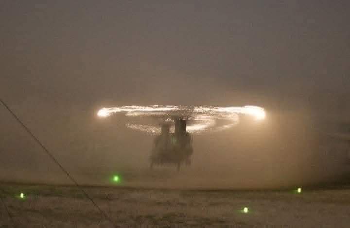 A helicopter is silhouetted against a dark, snowy night, its rotor blades glowing with bright lights that create a halo…