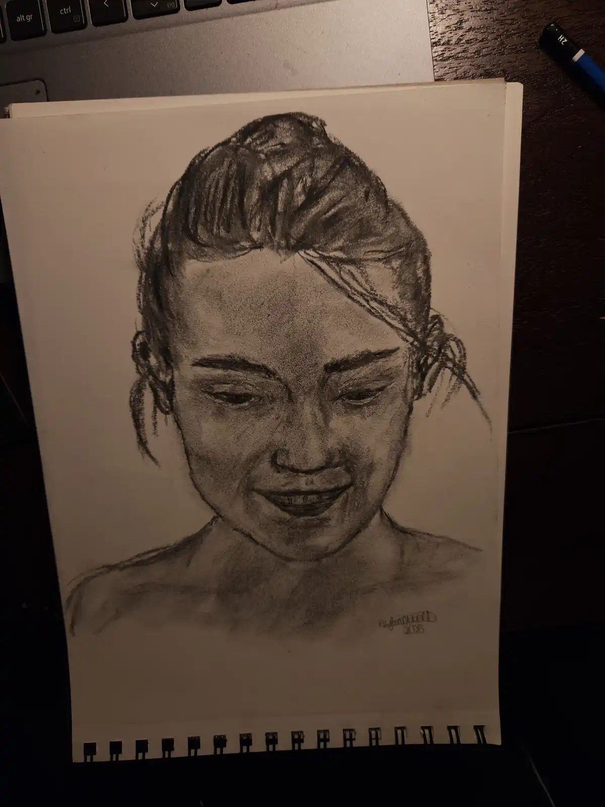 A charcoal sketch on spiral-bound paper shows a woman with her hair up, looking down with a tearful, smiling expression. The…