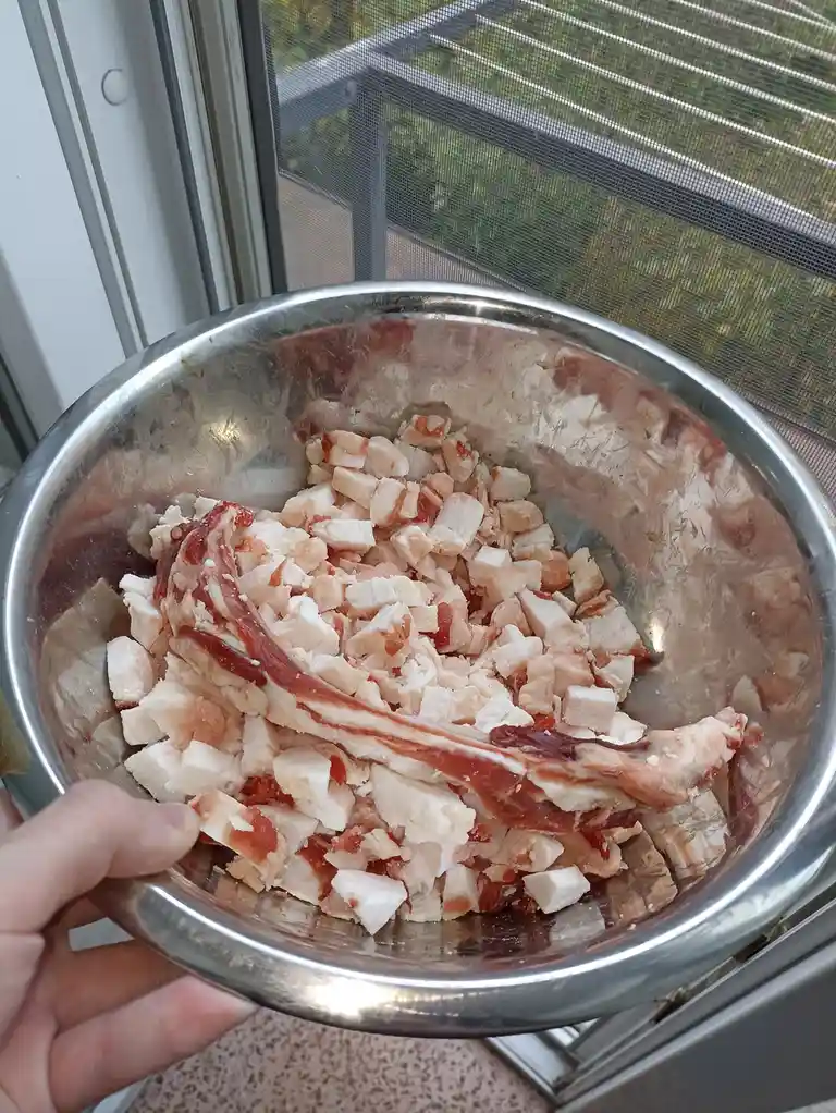 A hand holds a stainless steel bowl filled with chopped raw bacon pieces and one whole unchopped bacon strip. The bowl is…