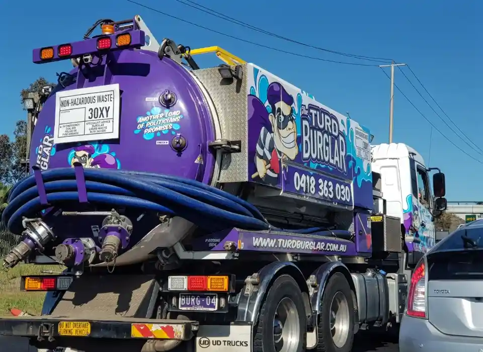A purple tanker truck labeled 'The Turd Burglar' with cartoon burglar graphics and contact info, has a white sign on its…